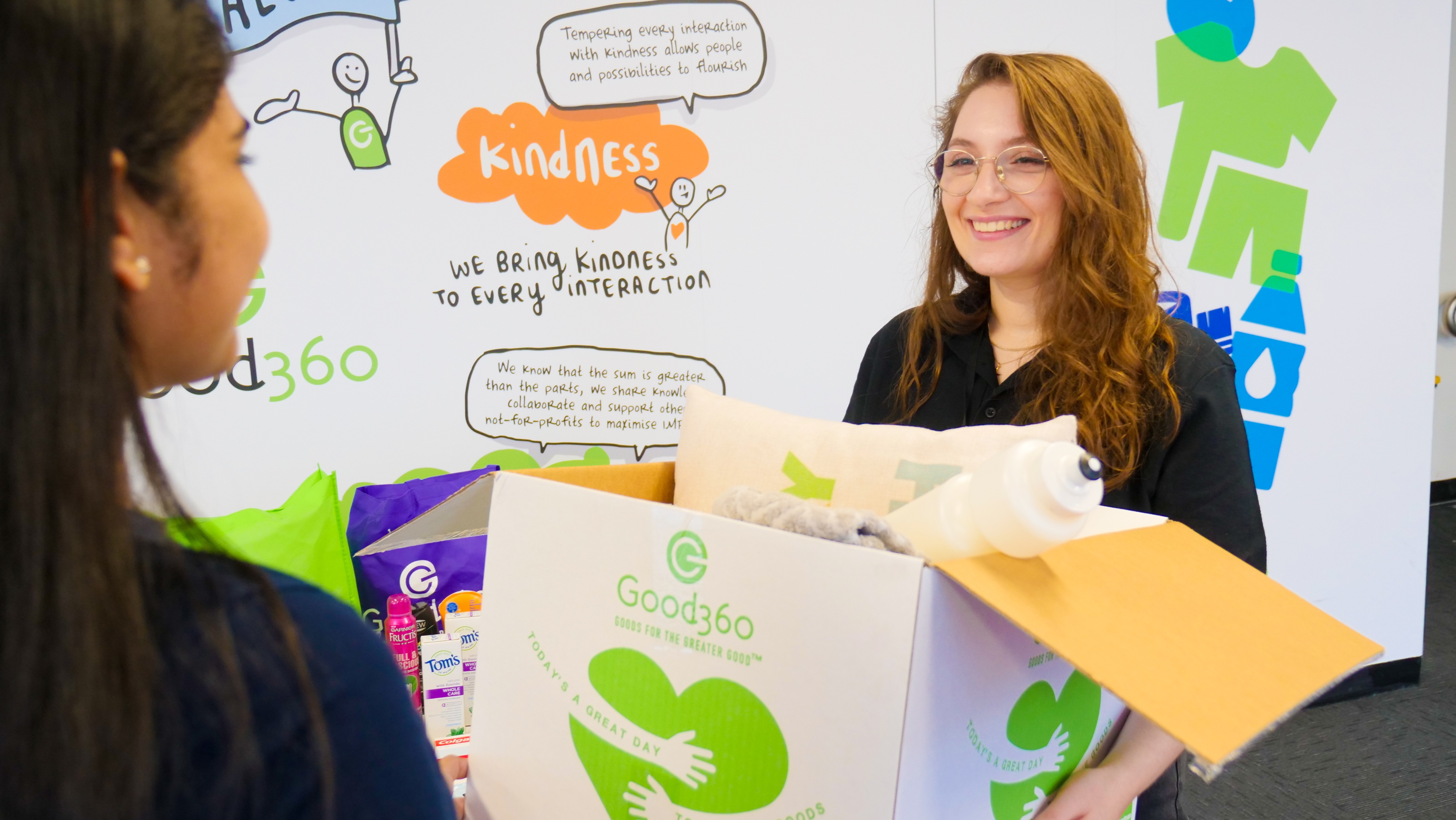 Smiling Good360 team member hands over a box of donated essential goods in a kindness-themed workspace.